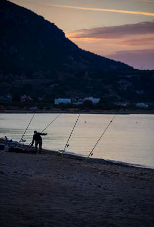 Silhouette of fisherman with fishing rod on the beach at sunsetの写真素材