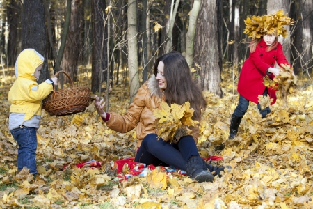 Mother with two children sits on an edge of the autumn wood, collecting leaves  The girl in a red coat jealously looks at the brotherの写真素材