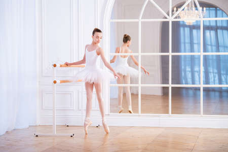 young ballerina stands on pointe shoes at a ballet barre in a beautiful white hall in front of a mirror.の写真素材