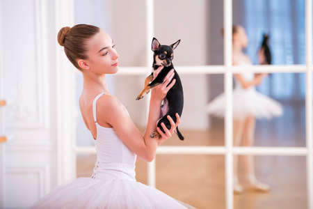 Young slender ballerina in a white tutu doing pointe shoes at a ballet bar in a beautiful white hall in front of a mirror.の写真素材