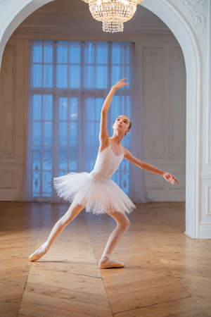 Young slender ballerina in a white tutu doing pointe shoes at a ballet bar in a beautiful white hall in front of a mirror.の写真素材