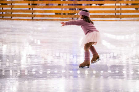 Little girl figure skater in a pink sweater is skating on a winter evening at an outdoor ice rink, back viewの写真素材