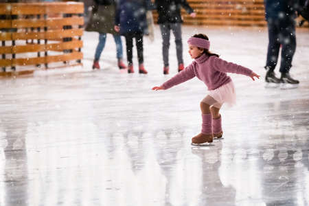 Little girl figure skater in a pink sweater is skating on a winter evening on an outdoor ice rink lit by garlandsの写真素材