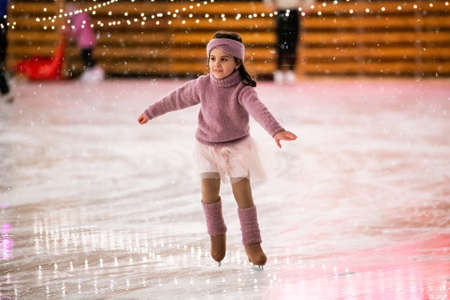 Little girl figure skater in a pink sweater is skating on a winter evening on an outdoor ice rink lit by garlandsの写真素材