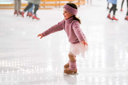 little girl in a pink sweater is skating on a winter evening on an outdoor ice rink lit by garlandsの写真素材