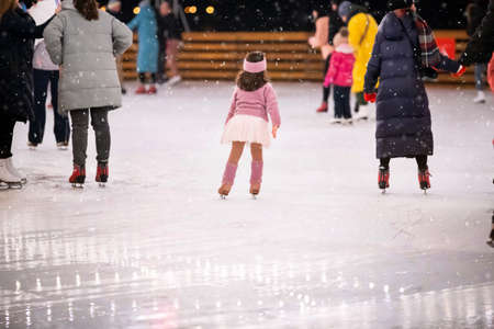 A little girl figure skater in a pink sweater rides on a winter evening on an outdoor ice rink among adults, rear viewの写真素材