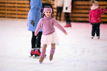 little girl in a pink sweater and a skirt is skating on a winter evening on an outdoor ice rink lit with garlandsの写真素材