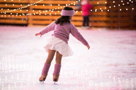 Little girl figure skater in a pink sweater is skating on a winter evening at an outdoor ice rink, back viewの写真素材