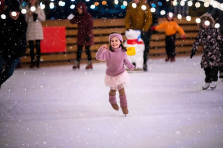 little girl in a pink sweater and a skirt is skating on a winter evening on an outdoor ice rink lit with garlandsの写真素材