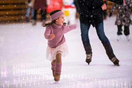Laughing little girl in a pink sweater is skating on a winter evening on an outdoor ice rink, it is snowingの写真素材