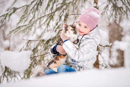 little girl sits with cat in an embrace in the snow in the winter forestの写真素材