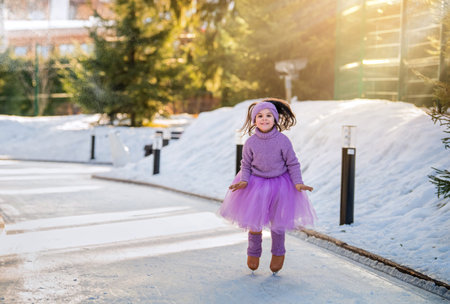 little girl in a pink sweater and a full skirt rides on a sunny winter day on an outdoor ice rink in the parkの写真素材