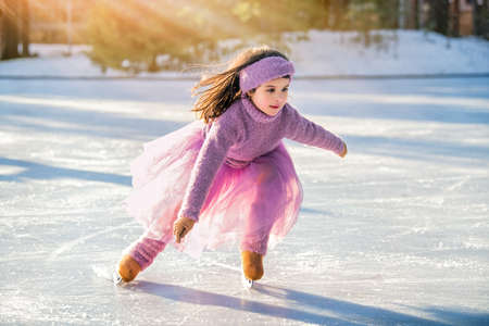 little girl in a pink sweater and a full skirt rides on a sunny winter day on an outdoor ice rink in the parkの写真素材