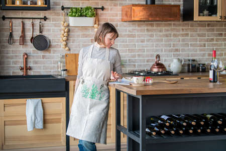 happy woman in a linen apron standing in the kitchen at the table examines a magazine.の写真素材