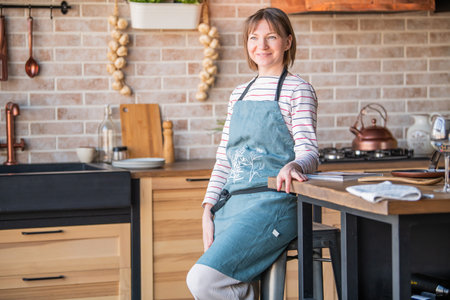 Happy woman in a linen apron standing in the kitchen at the table with a smile looks out the windowの写真素材