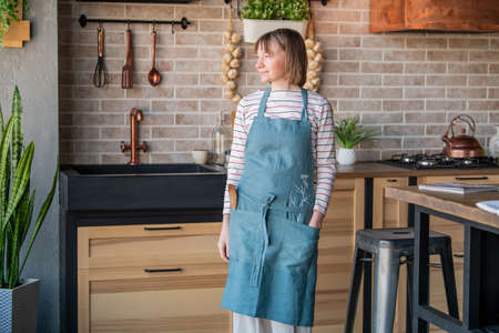 Happy woman in a linen apron standing in the kitchen at the table with a smile looks out the windowの写真素材