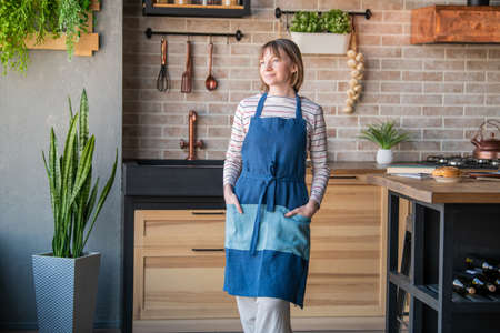 Happy woman in a linen apron standing in the kitchen at the table with a smile looks out the windowの写真素材