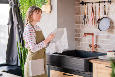 Happy woman in a linen apron standing in the kitchen by the sink wiping a plate with a linen towelの写真素材