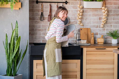 Laughing woman in a linen apron standing in the kitchen at the sink wiping a plate with a linen towelの写真素材