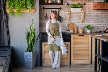 Happy woman in a linen apron standing in the kitchen by the sink wiping a plate with a linen towelの写真素材