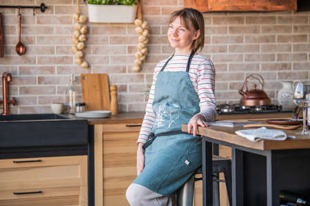 Happy woman in a linen apron standing in the kitchen at the table with a smile looks out the windowの写真素材