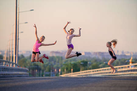 Three slender sports girls jumping on the road on a summer evening against the backdrop of the city panorama and pink sunset skyの写真素材