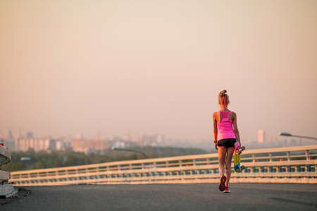 girl learns to ride a skateboard on the background of the evening city panoramas. leaves with a skateboard in his hands towards the cityの写真素材
