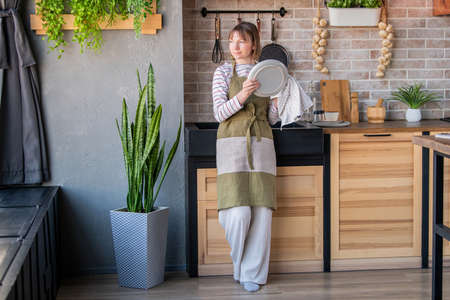 Happy woman in a linen apron standing in the kitchen by the sink wiping a plate with a linen towelの写真素材