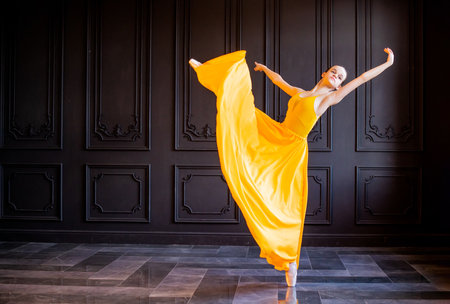 An elegant ballerina in pointe shoes dances with a flowing yellow fabric on a dark gray backgroundの写真素材