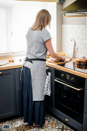 Happy woman in a linen apron stands in the kitchen next to the stove and cuts breadの写真素材