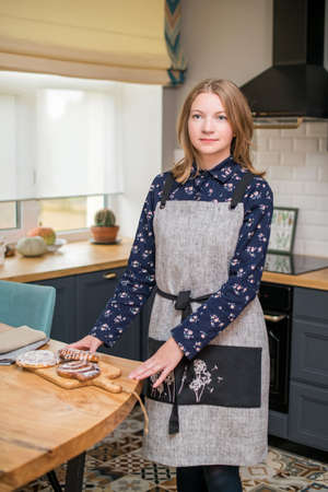 Happy woman in a linen apron stands in the kitchen at the table on which buns lieの写真素材