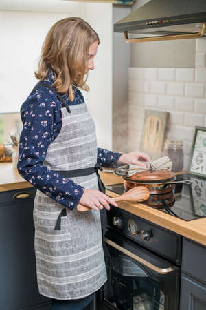Happy woman in a linen apron stands in the kitchen next to the stove and prepares foodの写真素材