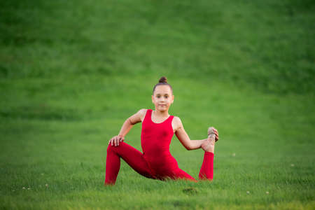 schoolgirl girl in a bright red overalls is engaged in gymnastics on the grass in the park, doing...
