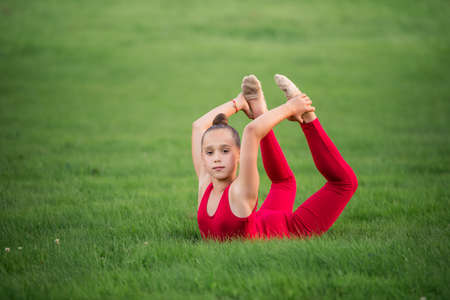 schoolgirl girl in a bright red overalls is engaged in gymnastics on the grass in the park, doing exercises for back flexibilityの写真素材