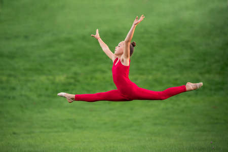 schoolgirl girl in a bright red overalls is engaged in gymnastics on the grass in the park, flies in a split jumpの写真素材