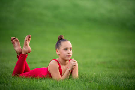 Girl schoolgirl gymnast in a bright red overalls is engaged in resting on the grass in the park, sniffing a clover flowerの写真素材