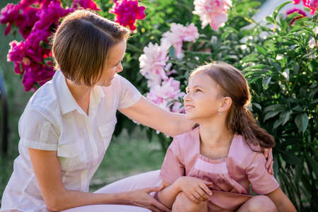 Happy family relationships. Mom and daughter sit on a bench and chat in the garden among the flowers on a sunny summer evening.の写真素材