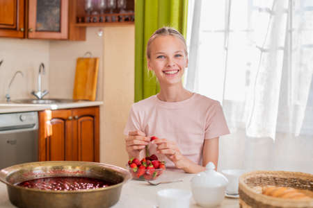 A girl at home in the kitchen sits at a table, eats strawberries, cooks strawberry jam, smiles a perfect smileの写真素材