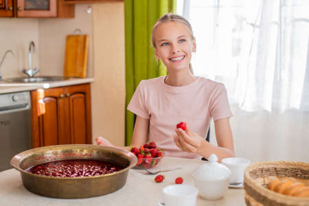 A girl at home in the kitchen sits at a table, eats strawberries, cooks strawberry jam, smiles a perfect smileの写真素材