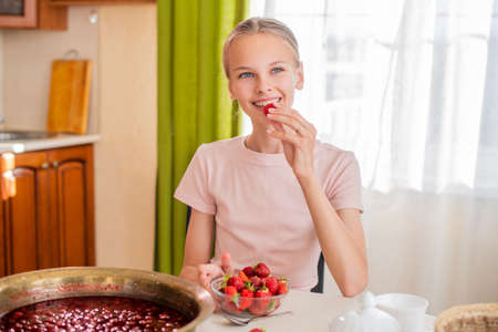 A girl at home in the kitchen sits at a table, eats strawberries, cooks strawberry jam, smiles a perfect smileの写真素材