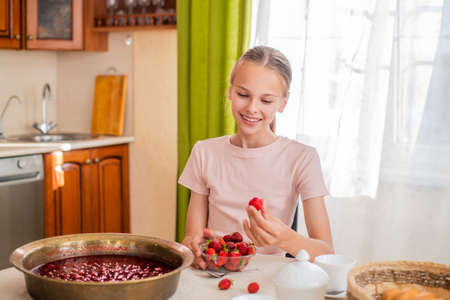 A girl at home in the kitchen sits at a table, eats strawberries, cooks strawberry jam, smiles a perfect smileの写真素材