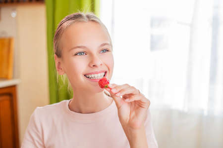 Teen girl eats strawberries at home in the kitchen, perfect teeth, charming smile, blue eyes. Close-up portrait.の写真素材