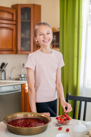 A girl at home in the kitchen stands near the table, eats strawberries, prepares strawberry jam, smiles with a perfect smileの写真素材