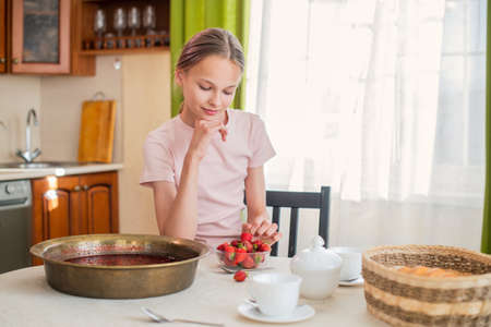 A girl at home in the kitchen sits at a table, eats strawberries, cooks strawberry jam, smiles a perfect smileの写真素材