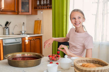 A girl at home in the kitchen sits at a table, eats strawberries, cooks strawberry jam, smiles a perfect smileの写真素材
