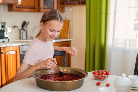 A girl at home in the kitchen sits at a table, eats strawberries, cooks strawberry jam, smiles a perfect smileの写真素材