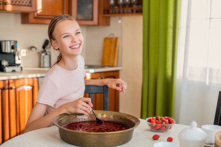 A girl at home in the kitchen sits at a table, eats strawberries, cooks strawberry jam, smiles a perfect smileの写真素材