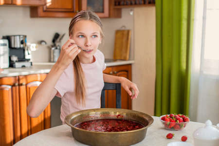 A girl at home in the kitchen sits at a table, eats strawberries, cooks strawberry jam, smiles a perfect smileの写真素材