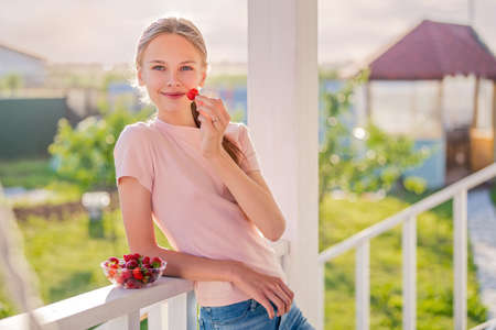 beautiful girl, blonde, teenager with long hair on a summer evening eats strawberries on the veranda of the cottageの写真素材