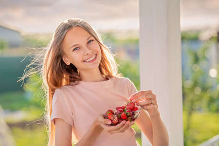 beautiful girl, blonde, teenager with long hair on a summer evening eats strawberries on the veranda of the cottageの写真素材